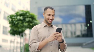 Gülümseyen genç bir yetişkin adam bir ofis binasının yanında caddede dururken akıllı telefon kullanıyor. Mutlu yakışıklı melez erkek e-postaları kontrol ediyor, internette sohbet ediyor, sosyal medyada geziniyor, mesajlaşıyor.