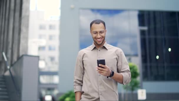 一位面带微笑的年轻人在一幢办公楼附近的街上散步时 用的是智能手机 快乐帅气的混血种族男性查电子邮件 网上聊天 浏览社交媒体 发短信 — 图库视频影像