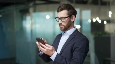 Resmi takım elbiseli yakışıklı işadamı, modern cam ofis sakallı, gözlüklü, göz gezdiren, sohbet eden akıllı telefon kullanan, iş yerinde kendine güvenen yatırımcı ya da aracı olan cep telefonu kullanıyor.