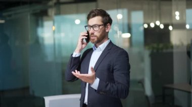 Kendine güvenen iş adamı yatırımcı modern ofisin içinde cep telefonuyla konuşuyor başarılı, meşgul, sakallı bir işçi iş yerinde akıllı telefondan konuşuyor.