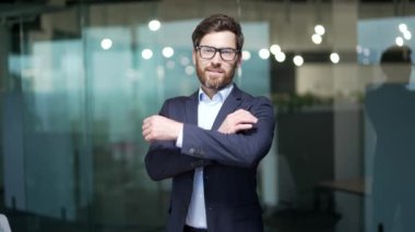 Başarılı yetişkin patronun portresi, gözlüklü ve iş adamı sakallı resmi takım elbiseli adam kameraya bakıyor ve gülümsüyor, kolları çaprazlı adam modern ofis binasında çalışıyor.