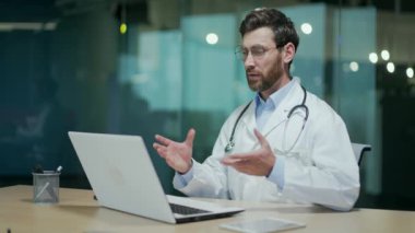 Steteskoplu kendine güvenen olgun doktor terapisti dizüstü bilgisayara bakıyor ve hastaya uzaktan kumandalı hastane konseptinde online video konferansında danışmanlık yapıyor.