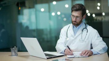 Steteskoplu kendinden emin olgun doktor terapisti dizüstü bilgisayara bakıyor, daktilo ediyor uzaktan kumandalı bilgisayarlara bakıyor ve modern hastanedeki hastanın tıbbi kartına not alıyor.