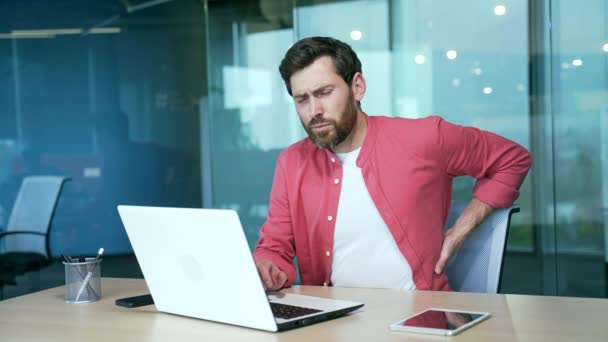 現代の職場のデスクでコンピューターのハードワークの後に激しい腰痛で疲れた髭のビジネスマンフリーランス悲しい起業家は室内オフィスで背骨の緊張に苦しんでいます — ストック動画
