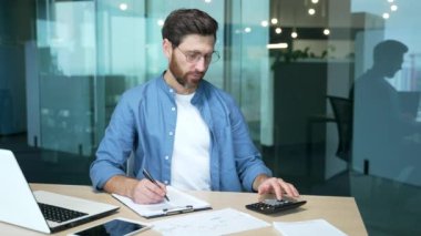 Odaklanmış iş adamı mali analist muhasebeci ya da evrak işleriyle uğraşan serbest muhasebeci hesap makinesindeki rapor grafiklerini sayıyor ve modern cam ofis işyerinde notlar alıyor.