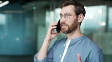 Kendine güvenen sakallı iş adamı gözlüklü girişimci modern ofisin içinde cep telefonuyla konuşuyor başarılı mutlu bir işçi iş yerinde akıllı telefondan profesyonel sohbetler yapıyor.