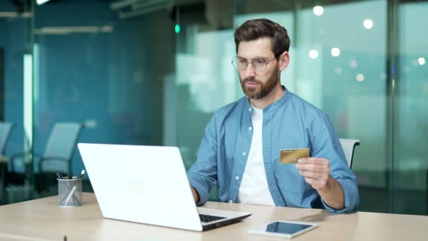 在现代化的办公室里 拿着信用卡坐在电脑前做银行付款的快乐人 男性企业家在办公桌前支付账单 在网上购物 欣喜若狂的商人赢了 — 图库视频影像