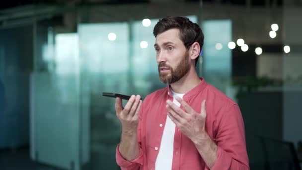 現代のオフィスの職場でスマートフォンを保持しながら ハンサムな成熟したビジネスマンフリーランスの男は声のメッセージを記録自信起業家は冷静にガジェットのマイクに屋内で話す — ストック動画