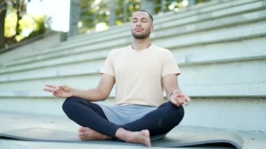 Genç bir sporcu, şehir stadyumundaki bir kilimde lotus pozunda meditasyon yapıyor. Eşofmanlı, melez, yakışıklı bir erkek sokakta yoga yapıyor. Sporcu gözleri kapalı gevşer