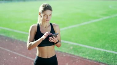 Yetişkin atletik kadın, şehir stadyumunda koşu bandında dururken akıllı telefondan antrenman sonuçlarını kontrol ediyor. Eşofman giyen fit bir kadın başarısından dolayı mutlu ve gülümsüyor ve evet işareti yapıyor.