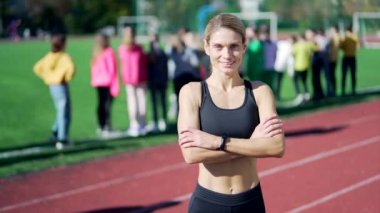 Şehir stadyumunda koşu bandında duran, eşofmanlı formalı formda bir sporcunun kollarını kavuşturup kameraya bakan, öğrencilerle mutlu bir antrenör portresi.