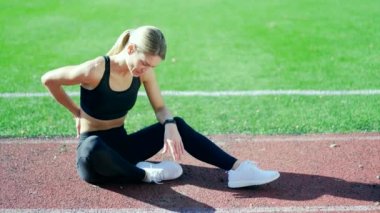 Olgun bir bayan koşucunun, şehir stadyumundaki koşu bandında oturan yaralı bir sırtı var. Kadın açık havada koşarken omurgasına zarar vermiş. Ağrıyı dindirmek umuduyla hastalıklı bölgeye masaj yapıyor.