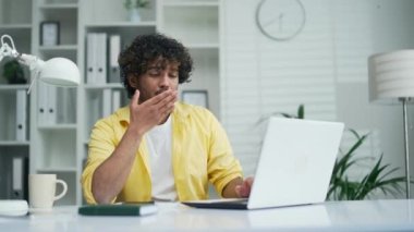 Uykucu, sıkkın adam, modern işyerindeki bir dizüstü bilgisayarın başında oturuyor. Erkek işçi esniyor, uyukluyor ve yorgun, üzgün, kıvırcık, serbest çalışan, sarı gömlekli bir uzman gibi görünüyor.