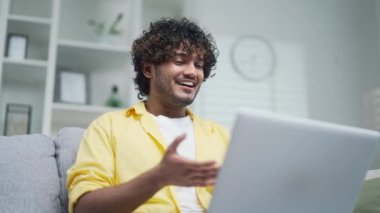 Neşeli adam, ailesi ya da arkadaşlarıyla bilgisayar kullanarak çevrimiçi iletişim kuruyor. Evde, kanepede oturan sarı gömlekli bir adam uzaktan konuşuyor, ellerini sallıyor ve gülümsüyor.