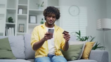 Genç yetişkin bir adam akıllı telefon ve kredi kartıyla ödeme yapıyor. Evde, kanepede oturuyor. Mutlu bir erkek telefonuyla mobil uygulama rezervasyonu yapıyor. İnternetten alışveriş yapıyor.