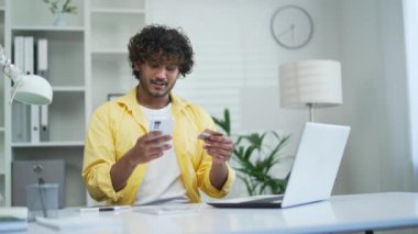 Genç yetişkin bir adam akıllı telefon ve kredi kartıyla ödeme yapıyor. Ofiste ya da evde oturuyor. Telefonla mobil uygulama rezervasyon siparişi veren mutlu bir erkek çevrimiçi mağazalardan alışveriş yapıyor.