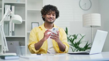 Sarı tişörtlü mutlu genç adam ev ofisteki telefonu kullanıyor. Karışık ırk erkek öğrenci daktilo kullanıyor. Uygulama sohbeti mesajları, kayan akıllı telefon gülümsemesi, kapalı alanda sörf yapan internet alışverişi yapıyor.