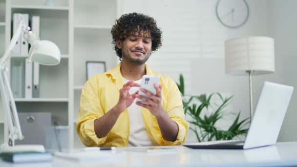 快乐英俊的年轻人穿着黄色衬衫在家里办公室用手机混合种族的男生打字浏览应用聊天短信滚动智能手机微笑室内冲浪网网上购物 — 图库视频影像