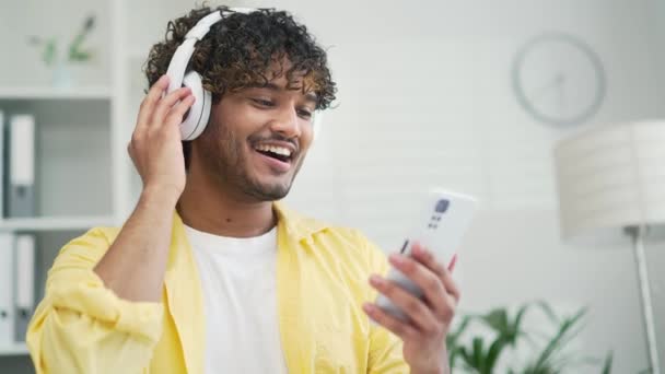 快乐的印度男性创业者 自由职业者或员工喜欢坐在室内 用智能手机应用办公软件或穿着黄色衬衫的学生来听耳机里的音乐 — 图库视频影像