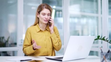 Olgun bir iş kadını müşterisiyle ya da iş arkadaşıyla telefonda konuşuyor. Ofiste bilgisayarın yanında oturuyor. Cam iş yerinde profesyonel sohbetler yapan meşgul sarışın bir kadın işçi.
