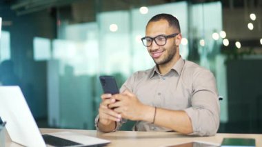 Gözlüklü genç bir işadamı modern bir ofisin çalışma masasında otururken akıllı telefon kullanıyor. Gülümseyen karışık ırk çalışanı sosyal medyada haberleri kontrol ediyor, internetten mesaj atıyor, e-postaları kontrol ediyor
