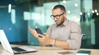 Modern ofisteki çalışma masasında otururken elinde akıllı telefon tutarak şok edici bir mesaja bakan genç işadamı. Karışık ırk adamı kötü haberleri okurken stresli hissediyor.