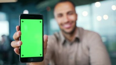 Modern ofiste oturan genç iş adamını gösteren yeşil ekranlı akıllı telefonu kapat. Renk anahtarı, reklam için dikey şablon düzeni. Gülümseyen melez adam başparmak işareti yapıyor.