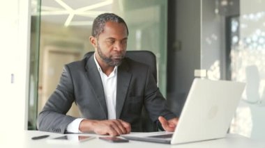 Ofiste dizüstü bilgisayar kullanarak resmi bir iş yerinde oturan kendinden emin Afro-Amerikan bir adamın portresi. Mutlu orta yaşlı işadamı dostça bir bakışla kameraya bakıyor.