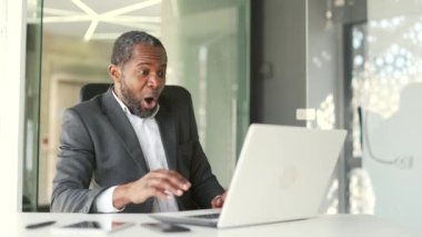 Heyecanlı, Afro-Amerikalı resmi takım elbiseli bir adam iş yerinde dizüstü bilgisayarını kullanarak spor maçı izlerken heyecanlı, duygusal jestleri olan favori takım için tezahürat yapan mutlu iş adamı.