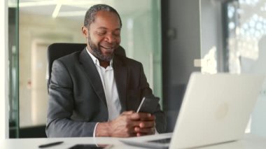 Modern ofisteki çalışma masasında otururken resmi takım elbiseli, gülümseyen Afro-Amerikan bir adam akıllı telefon kullanıyor. Mutlu orta yaşlı işadamı e-postaları kontrol ediyor, mesajlar yazıyor, internette sohbet ediyor.