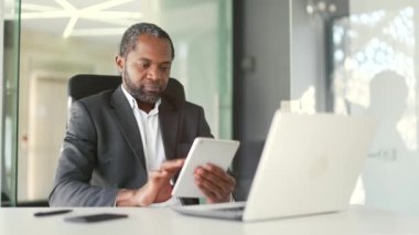 Modern ofisteki çalışma masasında otururken resmi takım elbiseli, konsantre olmuş Afro-Amerikan bir adam tablet kullanıyor. Gülümseyen orta yaşlı işadamı internette internetten bilgi topluyor