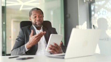 Resmi takım elbiseli, gülümseyen Afro-Amerikan bir adam modern ofiste iş yerinde otururken video ekranında tablet kullanarak konuşuyor. Çevrimiçi sohbet eden mutlu orta yaşlı iş adamı
