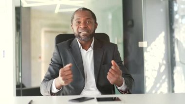 Webcam görüntüsü. Resmi takım elbiseli, gülümseyen Afro-Amerikan adam modern ofiste otururken kameraya bakıyor. Duygusal sohbeti olan kendine güvenen bir iş adamı.