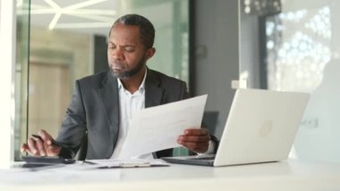 Resmi olarak odaklanmış Afro-Amerikan finansör elinde tuttuğu belgelerle modern ofiste çalışırken hesap makinesindeki verileri kontrol ediyor. Muhasebeci bir mali raporu analiz eder