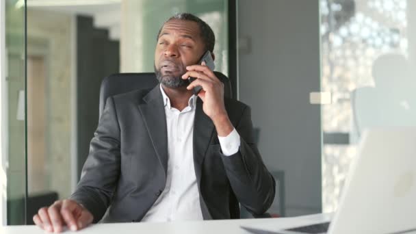 現代のオフィスのデスクで職場に座っている間 スマートフォンで話す正式な訴訟で成熟したアフリカ系アメリカ人男性を確信しています 重大なビジネスマンはビジネスパートナーと会話をしている — ストック動画