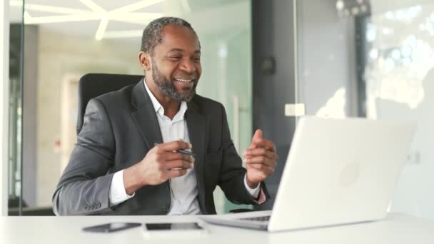 穿着正式西装的成熟的非洲裔美国人坐在现代办公室的工作场所 一边用笔记本电脑对着视频通话一边笑 快乐的中年商人在网上谈情说爱 — 图库视频影像