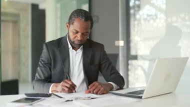 Resmi takım elbiseli kendinden emin Afro-Amerikan iş adamı, modern bir ofiste otururken hesap makinesiyle bir kağıt parçasına yazı yazıyor. Muhasebeci projenin maliyetini hesaplar.