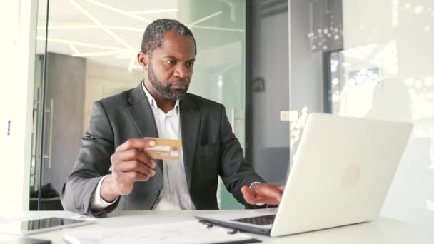 穿着正式西装的快乐的非洲裔美国人坐在办公室工作时 在笔记本电脑上输入信用卡号码 商人正在网上购物 对产品的高价格感到满意 — 图库视频影像
