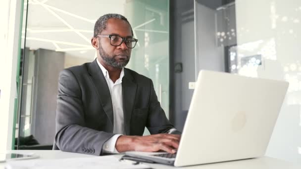 一位穿着西装和眼镜的严肃的非洲裔美国商人坐在现代化的办公室里 正在笔记本电脑上工作 一个信心十足的投资者会密切监察证券市场的情况 — 图库视频影像