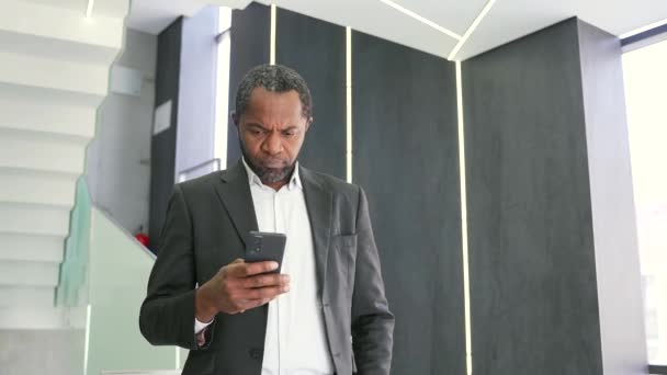 深刻なアフリカ系アメリカ人ビジネスマンがオフィスビル内に立っている間 スマートフォンを使って電子メールメッセージをチェックしています 思慮深い投資家は 証券取引所の状況を分析する — ストック動画