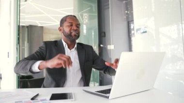 Resmi takım elbiseli olgun bir Afro-Amerikan erkeği modern bir ofiste masa başında otururken ellerini uzatarak rahatlıyor. İşadamı dinleniyor, işe ara veriyor.