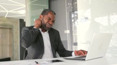 Modern ofisin çalışma masasında otururken boyun kası ağrısından muzdarip resmi takım elbiseli olgun Afro-Amerikan iş adamı. Orta yaşlı yorgun bir adam elleriyle hassas bir noktaya masaj yapıyor.