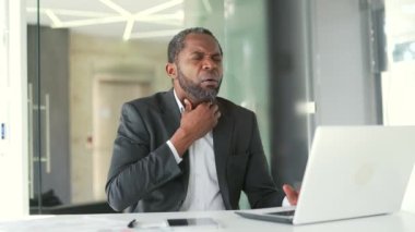 Resmi takım elbiseli olgun bir Afrikalı Amerikalı modern bir ofisin çalışma masasında otururken boğaz ağrısı çekiyor. Orta yaşlı üzgün bir erkek elleriyle bir yara yerine masaj yapıyor.