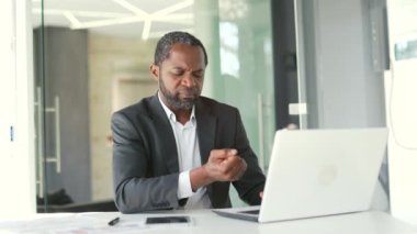 Modern ofiste çalışırken dizüstü bilgisayarda çalışırken bilek ağrısı çeken resmi takım elbiseli, olgun Afro-Amerikan iş adamı. Üzgün bir adam eliyle acı veren bir eklem masajı yapıyor.