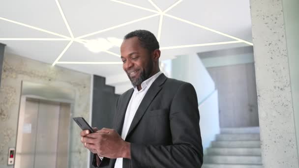 一位身穿正装 面带微笑的非洲裔美国商人站在一幢现代化的办公楼里看着智能手机 中年男子快乐发短信 上网与朋友聊天 — 图库视频影像