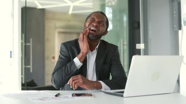 現代のオフィスの机に座っている間に歯痛に苦しむ正式な訴訟で成熟したアフリカ系アメリカ人男性 彼の手で頬をマッサージ悲しい中年の男性 歯医者の助けが必要だ — ストック動画