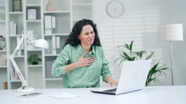 Kadın kalp krizi geçiriyor, hasta bir çalışan iş yerinde bilgisayarla çalışırken kalp ağrısı çekiyor. Takım elbiseli üzgün kadın elini içeride tutuyor.