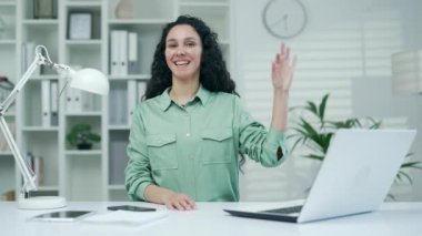 Kadın çalışan, kameraya bakarak iyi niyet gösteriyor. Tatmin olmuş, esmer, kadın çalışan, onay belgesi. İş yerindeki bilgisayar masasında oturuyor.