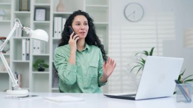 Genç, çekici Latin bir kadın iş yerinde modern bir ofiste otururken telefonda konuşuyor. Gülümseyen Kıvırcık Kıvırcık Esmer İş Kadını Kapalı Telefon Kullanarak İletişim