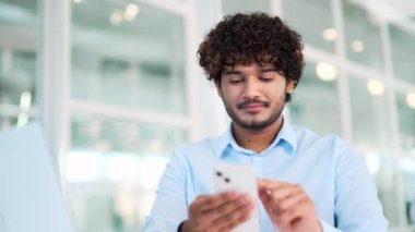 Kapatın. Genç yakışıklı bir işçi ofisteki bir çalışma masasında otururken akıllı telefon kullanıyor. Gülümseyen bir çalışan sosyal ağlara göz atıyor, çevrimiçi sohbet ediyor, daktilo ediyor veya mesaj okuyor